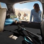A woman in blue stands by an open car trunk filled with hiking gear, overlooking a group walking in a sandy, mountainous area under a clear sky.