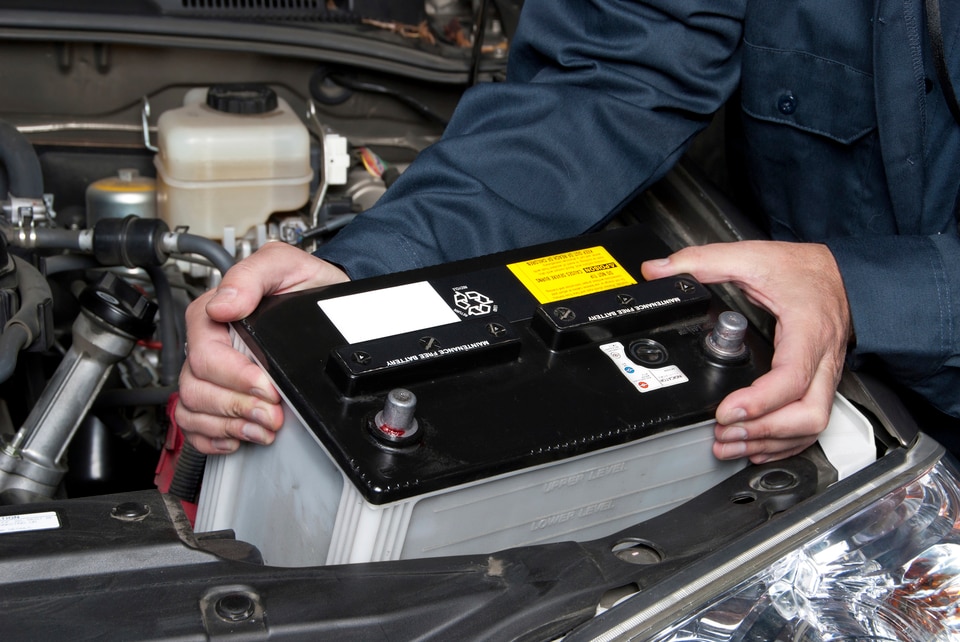 Mechanic installing a car battery under the hood. The battery is black with labels. The mechanic wears a blue uniform, conveying focus and precision.