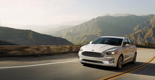 A silver sedan drives along a scenic mountain road under a clear sky. The sun casts a warm glow on rolling hills, conveying a sense of freedom and adventure.