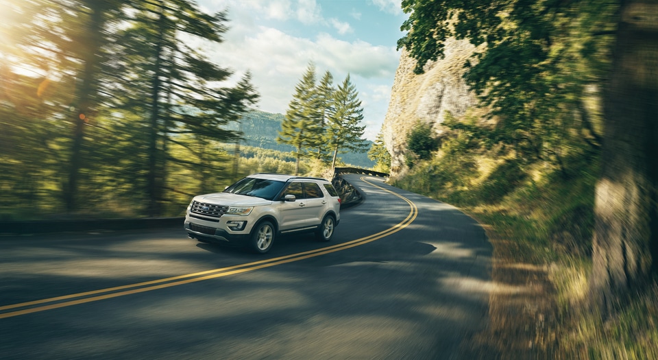 Silver SUV driving on a winding mountain road surrounded by lush greenery and rocky cliffs. The scene conveys adventure and freedom under a clear sky.