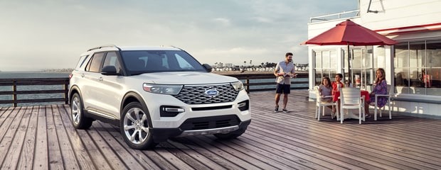 A white SUV is parked on a wooden pier beside a small cafe with a red umbrella. A family sits at a table, creating a relaxed, coastal atmosphere.