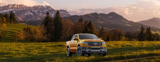 A yellow pickup truck is parked in a lush, green field with snow-capped mountains and tall trees in the background under a warm, dusk sky.