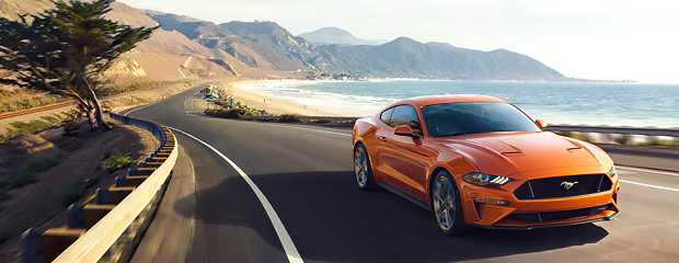 An orange sports car drives along a coastal highway with ocean waves and mountains in the background, conveying a sense of speed and freedom.