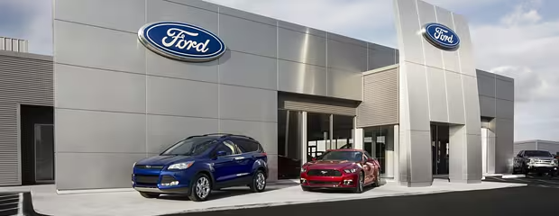 A modern Ford dealership with sleek architecture. A blue SUV and a red sports car are parked in front. The large Ford logo is prominently displayed.