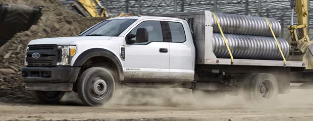 A white pickup truck navigates a dusty construction site, its flatbed loaded with large coiled pipes secured by yellow straps. Machinery is visible in the background.