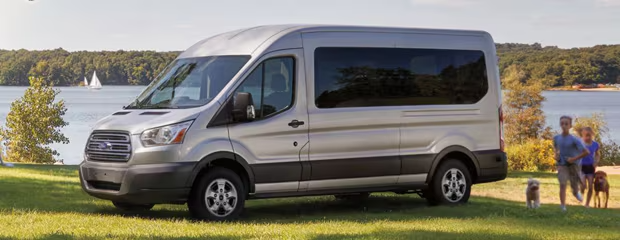 Silver van parked on grass near a lake, with a sailboat in the background. Two people and a dog walk on the right, conveying a relaxed, outdoor vibe.