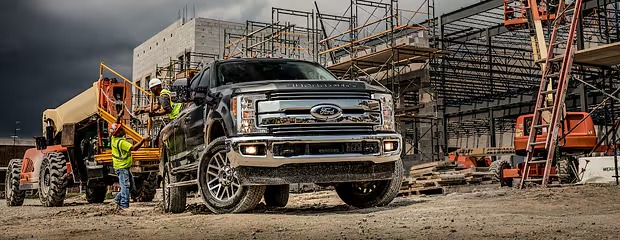 A black Ford truck is parked on a construction site surrounded by cranes and steel frameworks under a cloudy sky. Workers in safety gear are nearby, conveying a sense of industrious activity.
