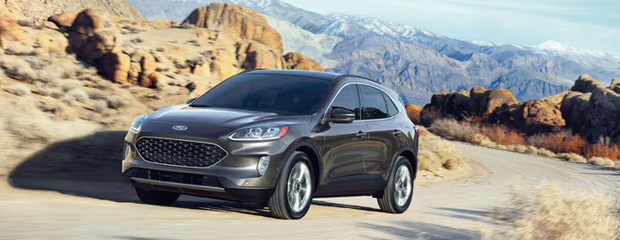 A gray SUV drives on a winding desert road, surrounded by rugged rocks and distant mountains under a clear blue sky. The scene conveys adventure.