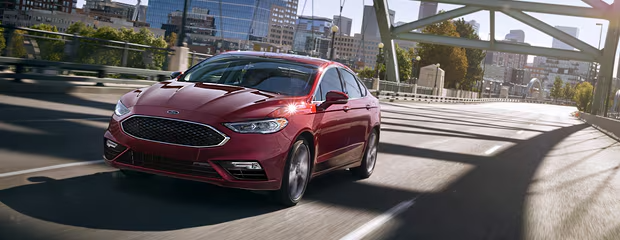 A sleek red sedan drives over a city bridge on a sunny day. The urban skyline and greenery are visible in the background, creating a dynamic, modern feel.