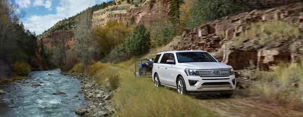 A white SUV towing a trailer drives alongside a scenic, tree-lined river with red rocky cliffs under a blue sky, conveying adventure and tranquility.