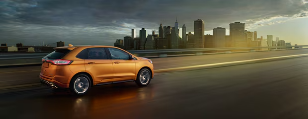 Orange SUV driving on a wet highway at dusk with a city skyline in the background. The scene is moody yet dynamic, suggesting motion and urban adventure.