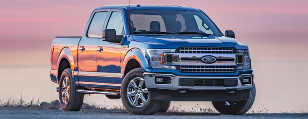 Blue Ford pickup truck parked on a gravel path at sunset. The sky is pink and orange, reflecting on the polished truck. Rugged and serene ambiance.