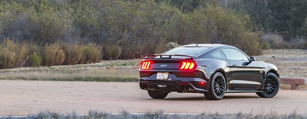 A sleek black sports car with illuminated rear lights is parked on an empty road. Surrounding it are green bushes, conveying a calm and serene setting.