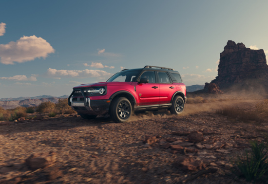 A red SUV drives through a rocky desert with dusty trails, under a clear blue sky and fluffy clouds, conveying adventure and ruggedness.