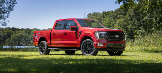 A red pickup truck is parked on a grassy field under clear skies. The background features lush green trees, suggesting a serene, outdoor setting.