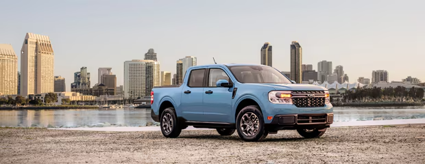 A light blue pickup truck is parked on a sandy surface with a city skyline in the background. The scene conveys a sense of adventure and urban exploration.