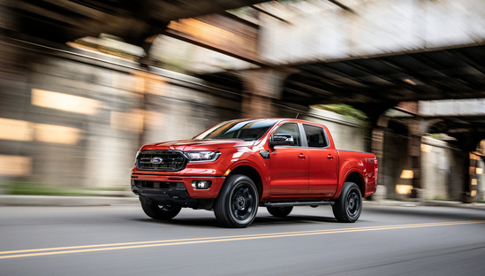 Red pickup truck driving on a city street under an old, rusted bridge. The motion blur conveys speed and a sense of adventure.