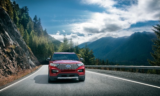 A red SUV drives on a winding mountain road, bordered by lush pine forests and rugged cliffs under a cloudy sky, evoking a sense of adventure.