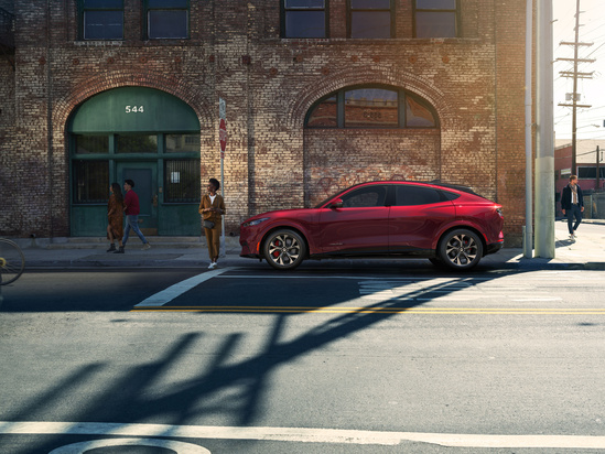 A sleek red SUV parked on a suburban street with modern houses and green trees in the background. A couple walks nearby, creating a calm, sunny scene.