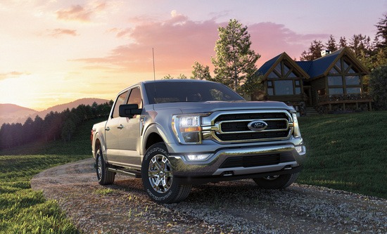 Silver pickup truck on a gravel path in front of a log cabin. Surrounded by lush greenery and a sunset sky, conveying a rustic, peaceful atmosphere.