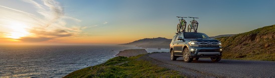 SUV with bicycles on roof drives along a coastal road at sunset, with cliffs and ocean on the left. The scene conveys adventure and tranquility.
