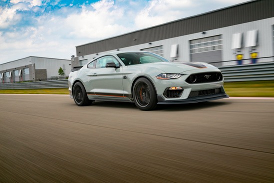 A gray sports car speeds down a racetrack beside industrial buildings, under a partly cloudy sky. The scene conveys speed and excitement.