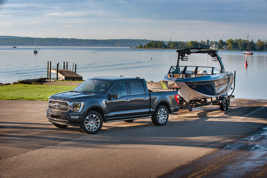 A dark gray pickup truck is parked on a lakeside road, towing a sleek boat. The serene lake and pier are in the background under a clear sky.