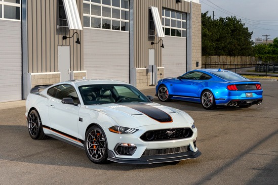 Two sporty Mustang cars, one white with black and orange accents, and one blue, parked in front of a gray industrial garage, conveying a sleek, powerful vibe.