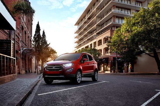 Red SUV parked on a quiet city street with modern buildings and lush trees. The scene is sunlit, conveying a warm, peaceful urban setting.