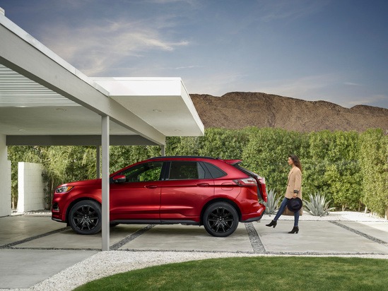 A red SUV is parked in a modern driveway with a mountain backdrop. A person in a tan coat and black pants approaches the car, holding a hat.