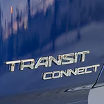 Close-up of a car's blue body with chrome lettering reading "Transit Connect." The surface is glossy, reflecting a distorted cityscape.