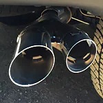 Close-up of a shiny black dual exhaust pipe on a vehicle, casting shadows on the ground. A tire with a distinctive tread pattern is visible nearby.