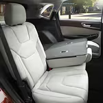 The car interior shows sleek, light gray leather seats with one backrest folded down, creating a cozy and modern aesthetic. Sunlight filters softly inside.