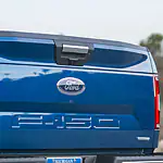 The image shows the tailgate of a blue Ford F-150 truck. The Ford logo and F-150 emblem are prominently displayed. The background is blurred.