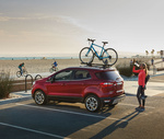 A red SUV with a bicycle on the roof parked by a beach. A person in athletic wear photographs the ocean. Beachgoers cycle in the background.