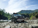 A rugged SUV is parked on a rocky riverbank with a backdrop of lush green forests and blue sky, conveying adventure and exploration.