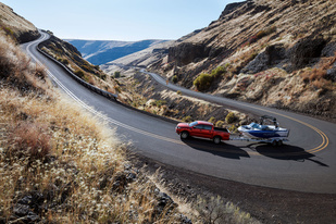 A red truck pulls a boat on a winding mountain road, surrounded by dry, grassy hills and a clear blue sky, evoking a sense of adventure and freedom.