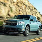 A light blue pickup truck driving on a scenic road with rocky cliffs in the background. The vehicle is in motion, conveying speed and adventure.