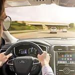A person with sunglasses drives a car on a sunny highway. The dashboard displays navigation apps. Two cars are ahead, with a bridge in the distance.