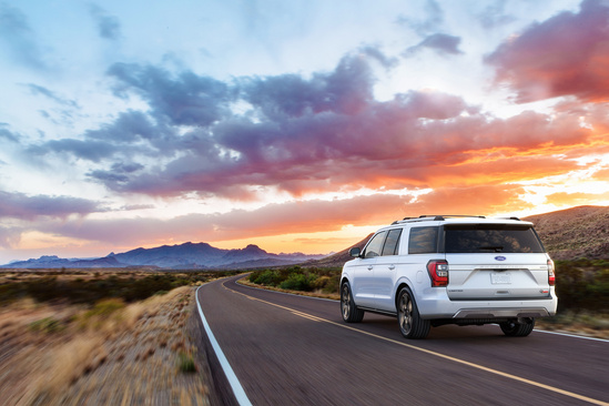 A white SUV drives on a deserted road through a desert landscape at sunset, with a dramatic sky of pink and orange clouds. The scene conveys adventure and tranquility.