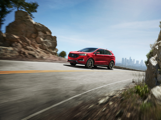 A red SUV speeds along a curving mountain road under a clear blue sky. Rocks line the road, with a city skyline visible in the distant background.