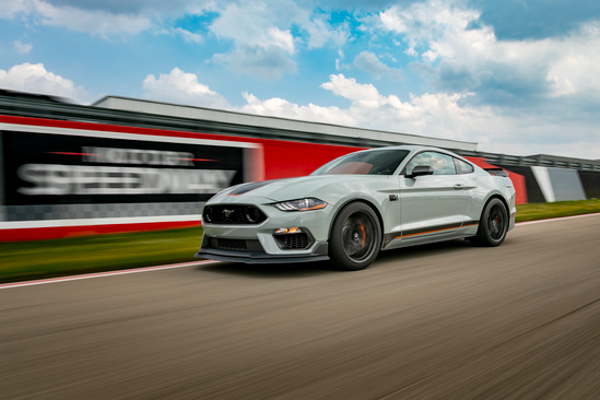 Sporty gray car racing on a track against a blurred background of a speedway, under a partly cloudy sky, conveying speed and excitement.