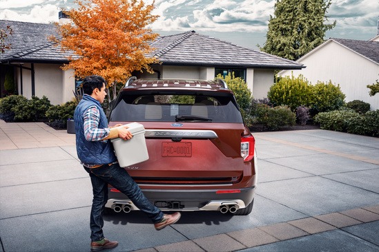 A person in a blue vest uses a kicking motion to open the trunk of a brown SUV parked in a driveway surrounded by greenery and autumn-colored trees.
