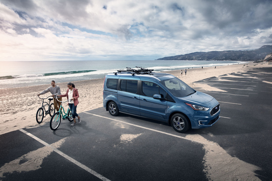 A blue van is parked in a beachside lot under a cloudy sky. Two people stand nearby with bicycles, conveying a relaxed, adventurous vibe.