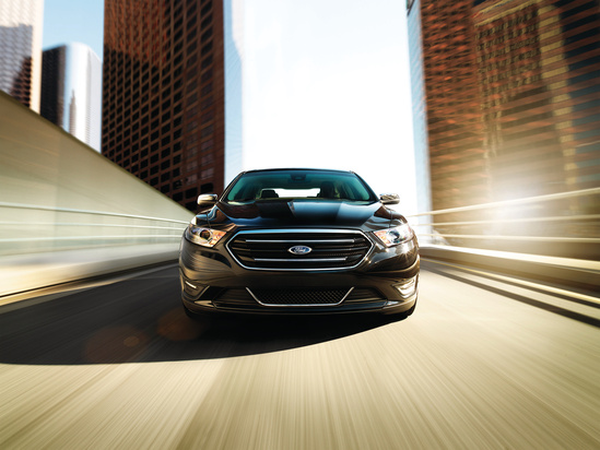 A black car drives quickly on a city bridge, creating a sense of speed. Tall buildings blur in the background under a bright sky, highlighting urban energy.