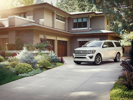 A white SUV is parked in the driveway of a modern brick house surrounded by lush greenery and colorful flowerbeds under a sunny sky.