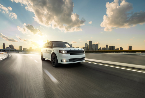 A white SUV speeds along an empty highway, with a vibrant city skyline and dramatic clouds at sunset, conveying freedom and adventure.