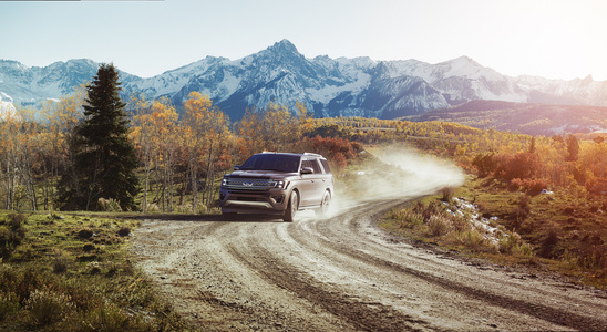 SUV driving on a winding dirt road through a scenic mountain landscape. Snow-capped peaks and autumn trees surround the vehicle, evoking adventure.