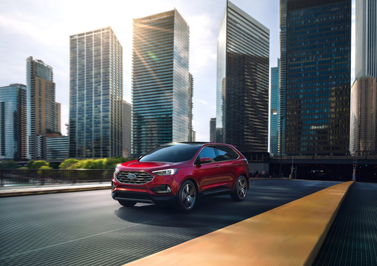 A red SUV drives along a city road flanked by tall, modern skyscrapers under a bright sky. The scene conveys a sense of urban sophistication and motion.
